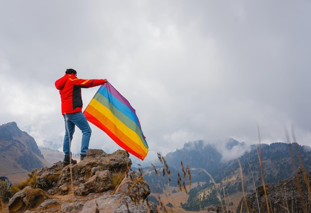 Hombre de pie en la cima de la colina y sosteniendo la bandera del orgullo LGBT