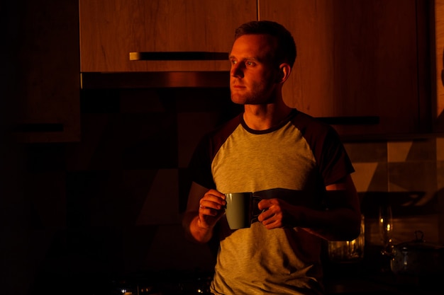 Un hombre de pie delante de una ventana en la iluminación del atardecer está bebiendo café en su cocina