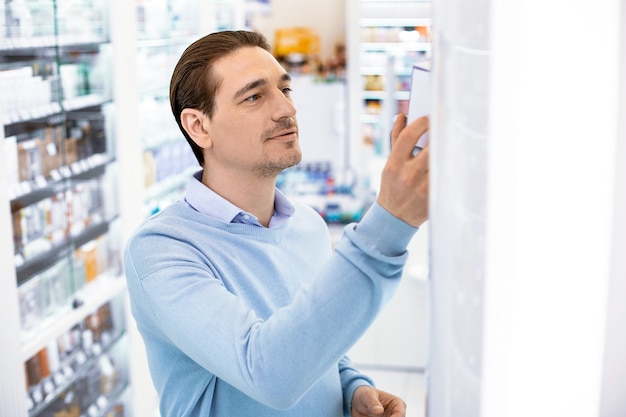 Un hombre de pie en una farmacia frente a la ventana que muestra un medicamento del estante, sonriendo