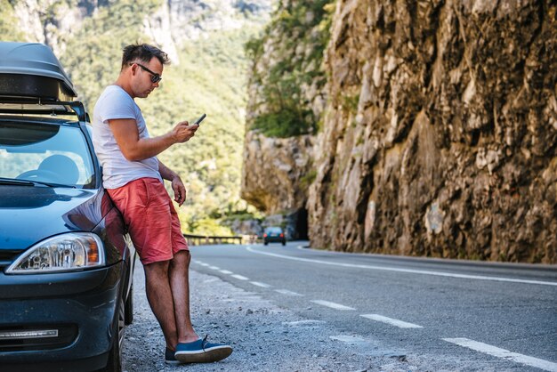 Hombre de pie junto al auto Broke Down y usando un teléfono inteligente