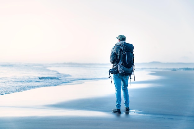 Hombre de pie en la orilla de la playa con mochila mirando el horizonte al atardecer