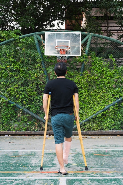 Foto hombre de pie con la pierna rota en el molde de yeso con muletas en la cancha de baloncesto