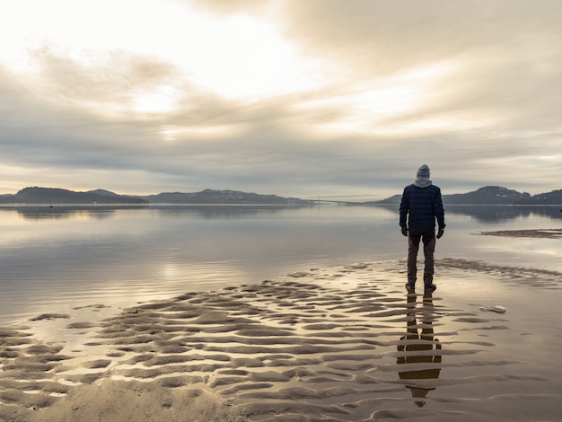 Hombre de pie en la playa, reflejos del hombre en el agua. Mar tranquilo, niebla y niebla. Hamresanden, Kristiansand, Noruega