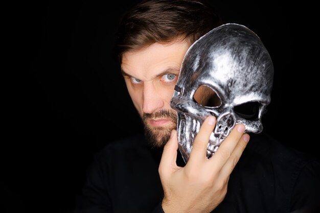 Un hombre de pie sobre un fondo negro mira desde debajo de una máscara de esqueleto que quiere ponerse en la cara