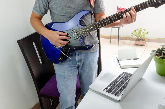 Hombre de pie tocando una guitarra eléctrica frente a una computadora en clases