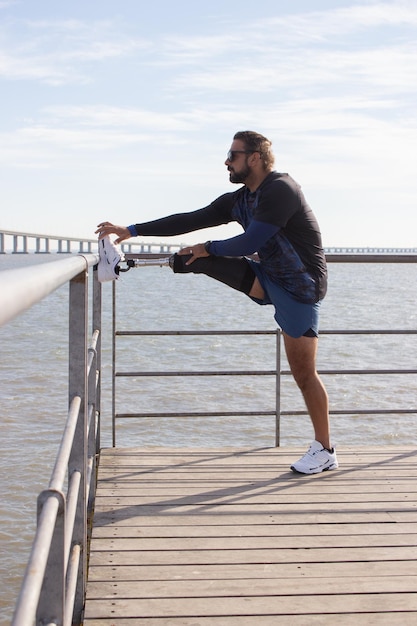 Hombre con pierna mecánica preparándose para entrenar en terraplén. Hombre con ropa deportiva estirando la pierna en el parque el día de verano. Deporte, concepto de entrenamiento