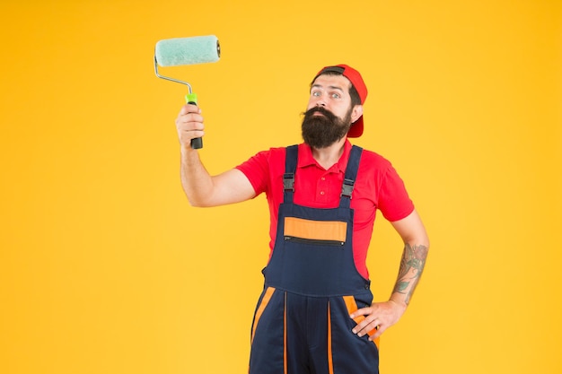 El hombre pinta el constructor de hipster de pared con gorra amarilla asistente de montaje de fondo en diseñador de ingeniero de uniforme de trabajo listo para pintar el concepto de reparación de pared trabajador de hombre barbudo con herramienta de rodillo