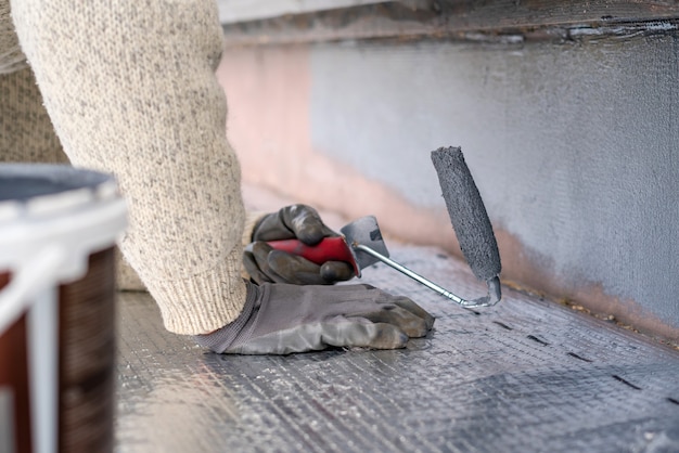 Hombre pintando casa zócalo con rodillo de pintura. Ciérrese para arriba de la pintura gris del balanceo en el zócalo del edificio. Renovación de viviendas