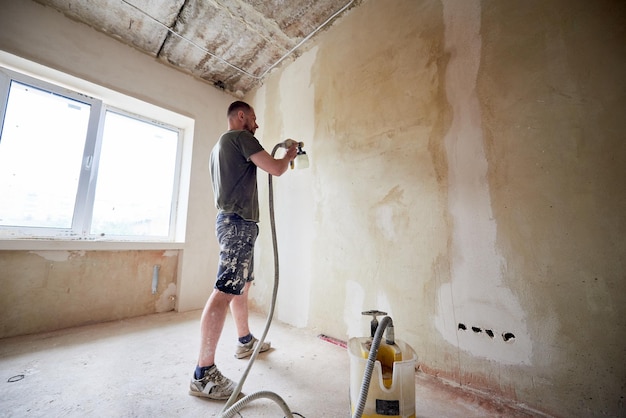 Hombre pintando una pared gris con una pistola rociadora renovando una casa nueva Recipiente abierto con pintura