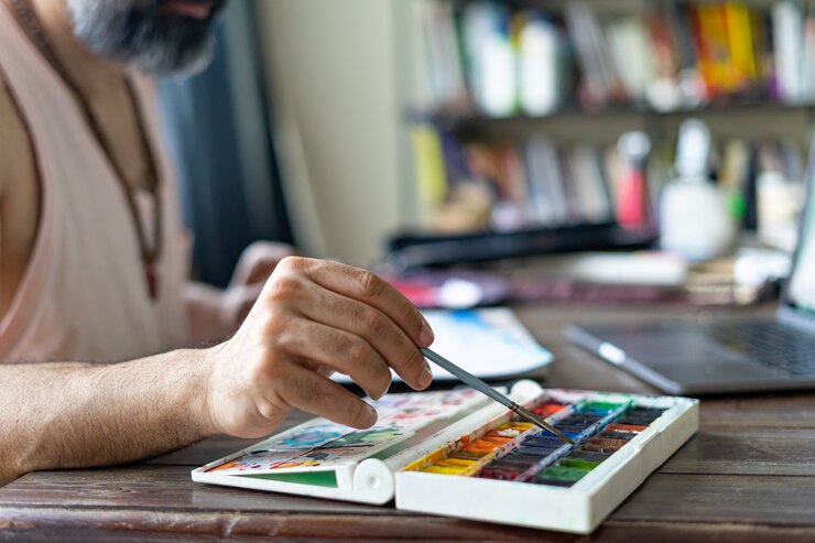 Foto hombre pintando con pincel y paleta de acuarela sobre papel delante de la computadora portátil