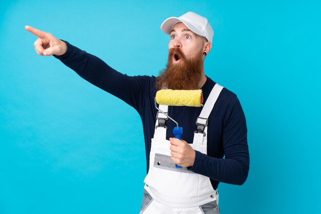 Hombre pintor con barba larga sobre pared azul aislado apuntando lejos