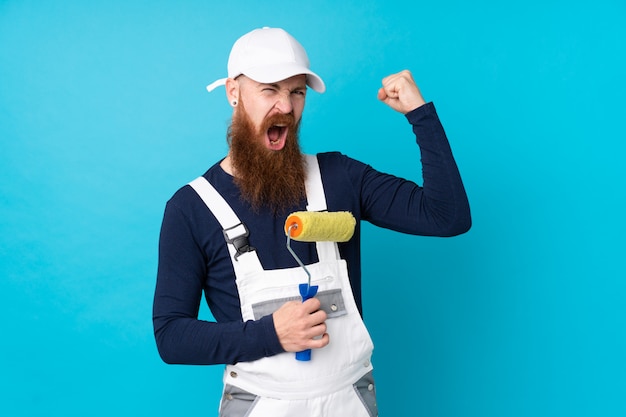 Hombre pintor con barba larga sobre pared azul haciendo un gesto fuerte