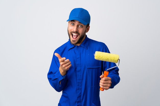 Hombre pintor sosteniendo un rodillo de pintura aislado en la pared blanca apuntando hacia el frente y sonriendo