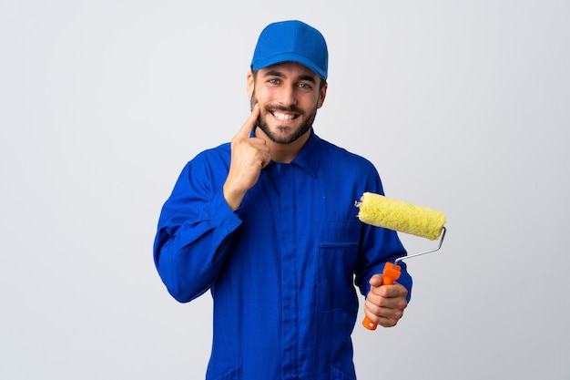 Hombre pintor sosteniendo un rodillo de pintura aislado en la pared blanca sonriendo con una expresión feliz y agradable