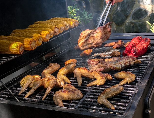 Hombre con pinzas de metal para convertir algunas alitas de pollo picantes que se asan a la parrilla en la barbacoa. Deliciosas alitas de carne de pollo asado y costillas de cerdo con mazorcas de maíz en la parrilla de barbacoa.