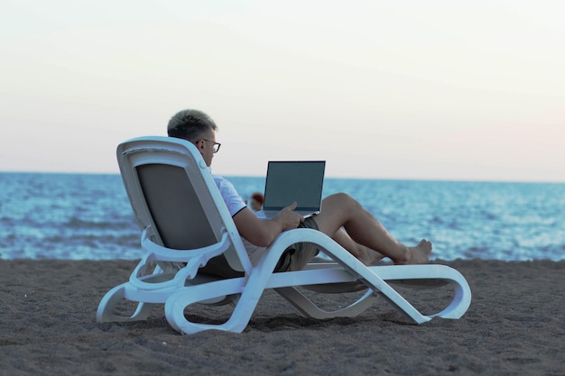 Un hombre en la playa con una computadora portátil en una silla de estar trabajando el concepto de trabajar en línea en cualquier lugar del mundo