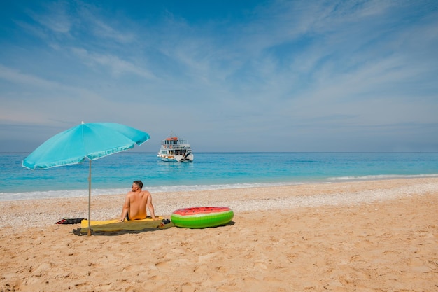 Hombre en la playa del mar día soleado sombrilla sombrilla Grecia vacaciones pequeño crucero en el fondo