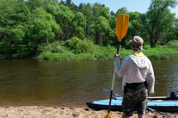 Un hombre se para en una playa con un remo y un kayak al fondo.