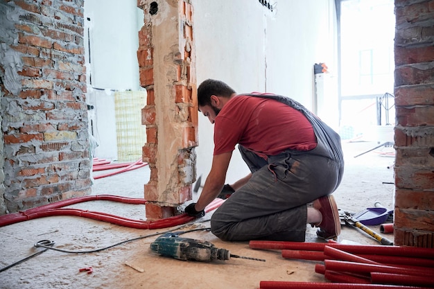 Hombre plomero en overoles de trabajo usando un martillo mientras coloca tuberías en el piso Trabajador instalando un sistema de calefacción mientras trabaja en la renovación del apartamento Concepto de renovación del hogar