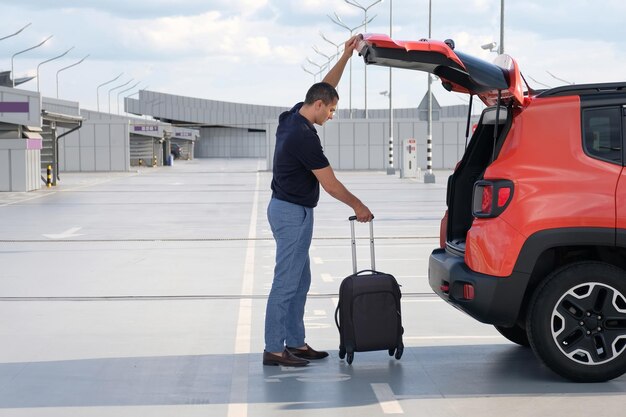 Un hombre pone su maleta en el maletero de un coche en un estacionamiento del aeropuerto concepto de vacaciones de viaje