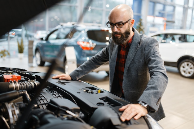 El hombre posa en la nueva camioneta con capó abierto, concesionario de automóviles. Cliente en la sala de exposición de vehículos, persona del sexo masculino que compra transporte, negocio de concesionario de automóviles