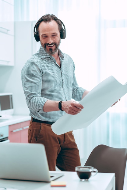 Hombre positivo con auriculares sonriendo a la pantalla del portátil mientras está de pie con un plano en la cocina