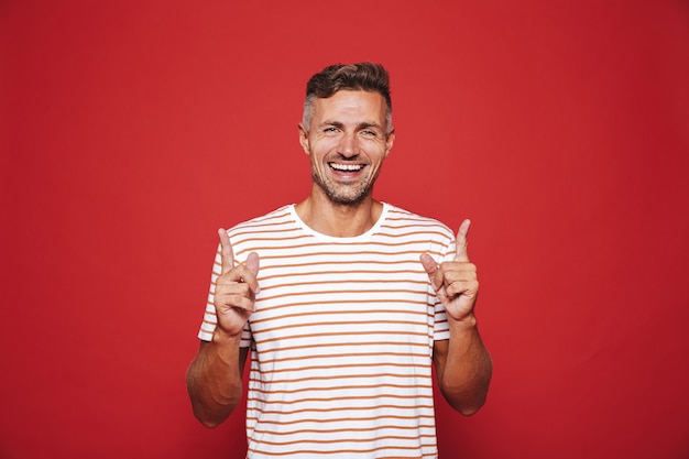 Hombre positivo en camiseta a rayas gesticulando los dedos índices hacia arriba en copyspace aislado en rojo