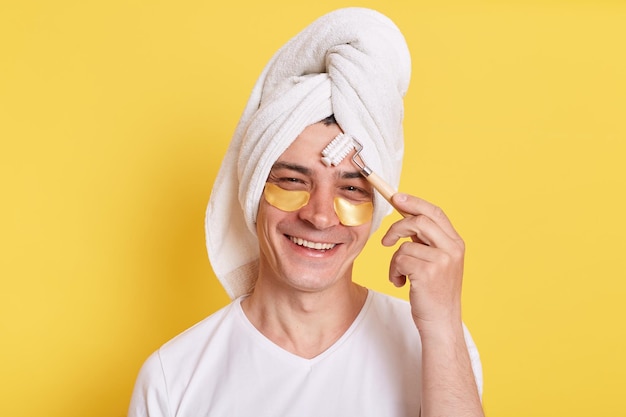 Un hombre positivo envolvió una toalla en la cabeza haciendo un masaje matutino para masajear las mejillas con parches debajo de los ojos, procedimientos de cosmetología aislados sobre fondo amarillo