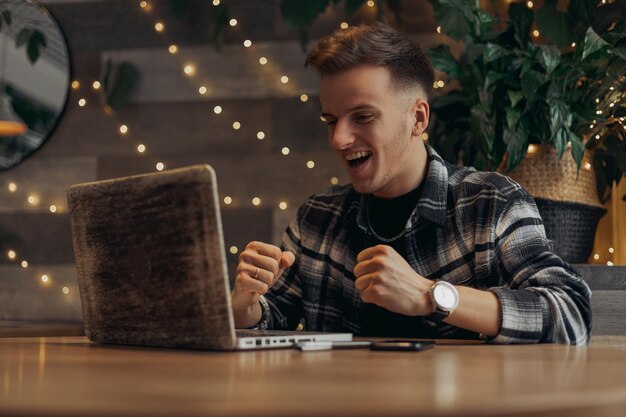 Hombre positivo leyendo buenas noticias en la computadora portátil y celebrando el éxito con los puños cerrados