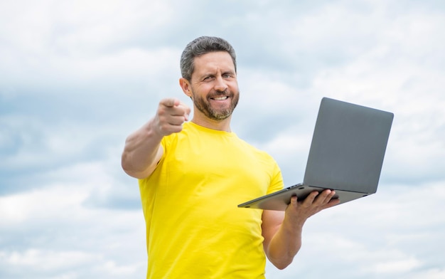 Hombre positivo que trabaja en línea en la computadora portátil en el fondo del cielo