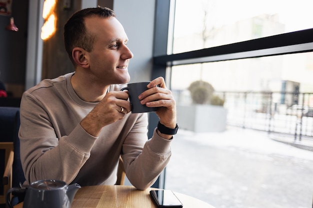 Un hombre positivo se sienta en una cafetería en una mesa, bebe café aromático. Un tipo sonriente con ropa informal se sienta en una cafetería y bebe café por la mañana.