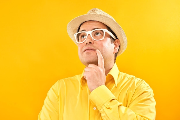El hombre positivo con sombrero de Panamá sosteniendo la barbilla piensa en las dudas de las vacaciones y toma una decisión aislada en el fondo amarillo del estudio