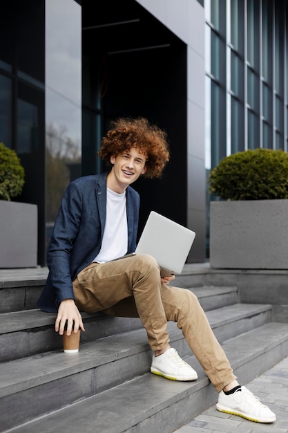 Hombre positivo sonriente sentado en las escaleras usando una computadora portátil sosteniendo una taza de papel con café