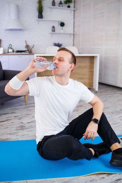 El hombre practica deportes en casa. Atleta bebe agua mientras está sentado en el suelo en casa, estilo de vida saludable