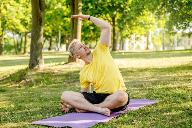 Hombre practicando yoga sentado en una alfombra al aire libre en verano Chico haciendo ejercicios de pilates para estirar los músculos