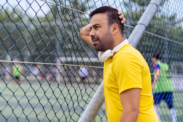 Hombre preocupado con auriculares inalámbricos viendo el entrenamiento del club de fútbol en un gran campo urbano