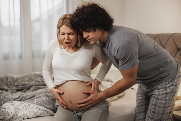 Hombre preocupado ayudando a su esposa embarazada en un dormitorio que está teniendo contracciones prenatales.