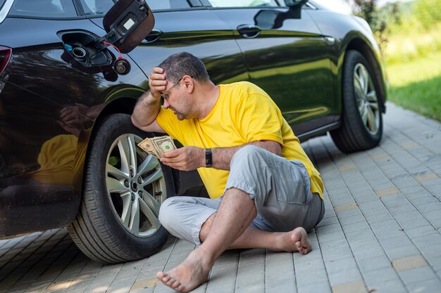 Hombre preocupado sentado en la acera junto a un automóvil cerca de un concepto de aumento de precio de combustible de tanque de combustible abierto