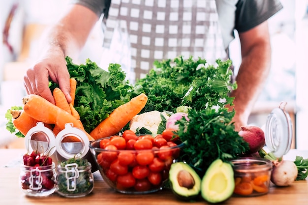 Hombre preparando comida vegetariana y vegana - mesa llena de verduras frescas y saludables de temporada - mezcla de colores y vitaminas - enfoque en la mano tomando una zanahoria - concepto de estilo de vida de bienestar