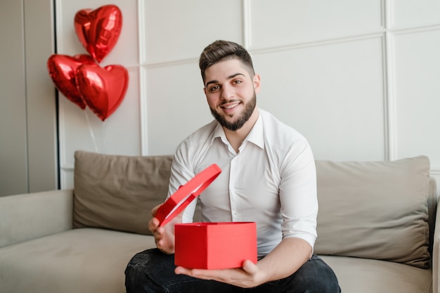 Hombre con presente en forma de corazón rojo en caja de regalo sentado en el sofá con globos en casa