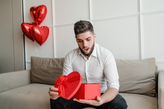 Hombre con presente en forma de corazón rojo en caja de regalo sentado en el sofá con globos en casa