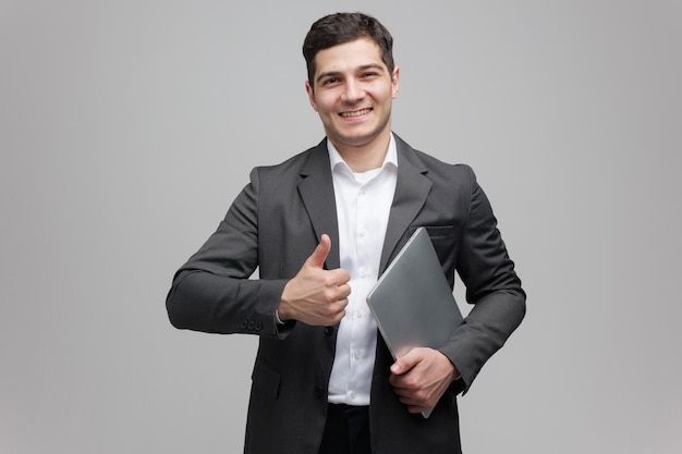 Hombre profesional en traje de negocios sonríe con confianza mientras sostiene una computadora portátil y levanta el pulgar