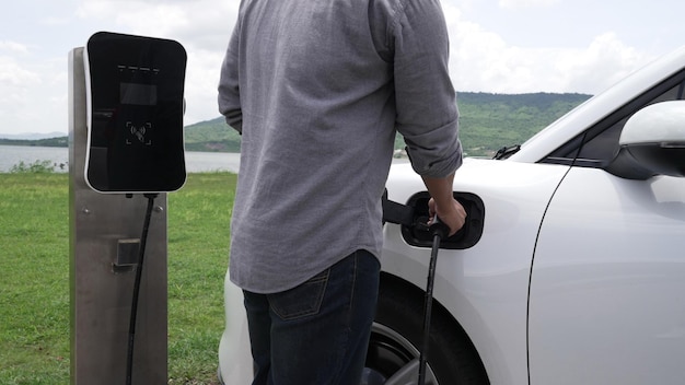 Un hombre progresista conecta el enchufe del cable de alimentación de la estación de carga a su automóvil eléctrico EV impulsado por energía verde en un campo verde para un futuro concepto de medio ambiente limpio