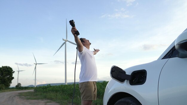 Foto hombre progresista con su coche eléctrico coche ev recargando energía desde la estación de carga en campo verde con turbina eólica como concepto de energía sostenible futura vehículo eléctrico con generador de energía