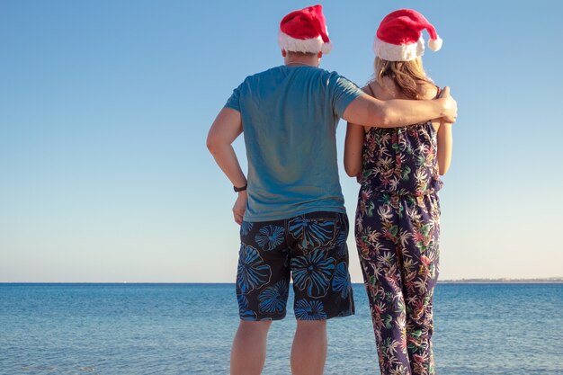 Hombre que abraza a la mujer en sombreros de la Navidad y que mira la playa del mar.