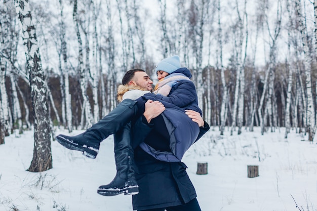 Hombre que llevaba a su novia en las manos en el bosque de invierno. Gente divirtiéndose al aire libre. Pareja relajante