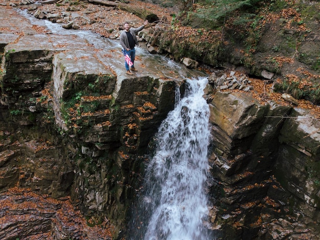 Hombre que sostiene la bandera de estados unidos en la parte superior de la cascada de otoño del acantilado