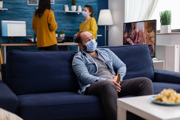 Hombre que sostiene una botella de cerveza relajándose en el sofá pasando tiempo con amigos manteniendo el distanciamiento social con mascarilla en la sala de estar mirando pensativo a la cámara evitando la propagación del coronavirus. Nueva pieza normal