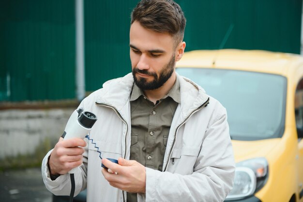 Hombre que sostiene el cable de carga de energía para el automóvil eléctrico en el estacionamiento al aire libre Y va a conectar el automóvil a la estación de carga en el estacionamiento cerca del centro comercial