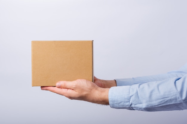 Hombre que sostiene la caja a distancia del brazo en la pared blanca. Caja cuadrada de cartón. Entrega de paquetes. Vista lateral.